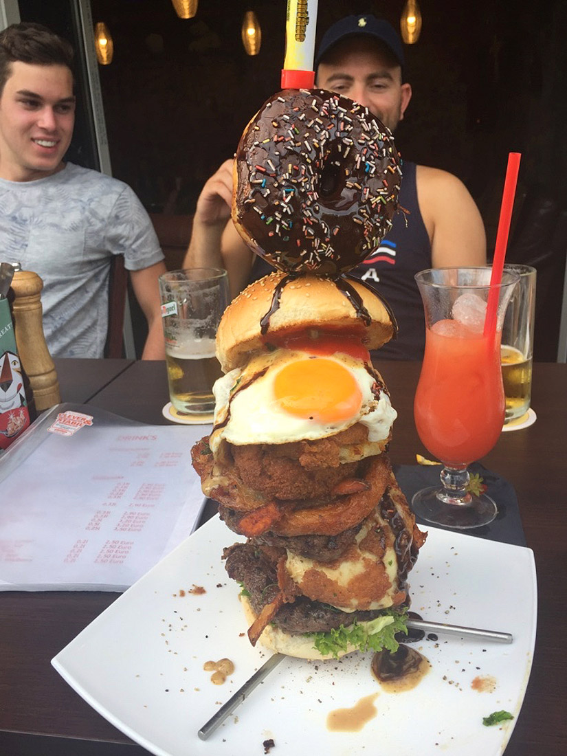 A photo of a larger-than-life burger, with a chocolate donut on top.