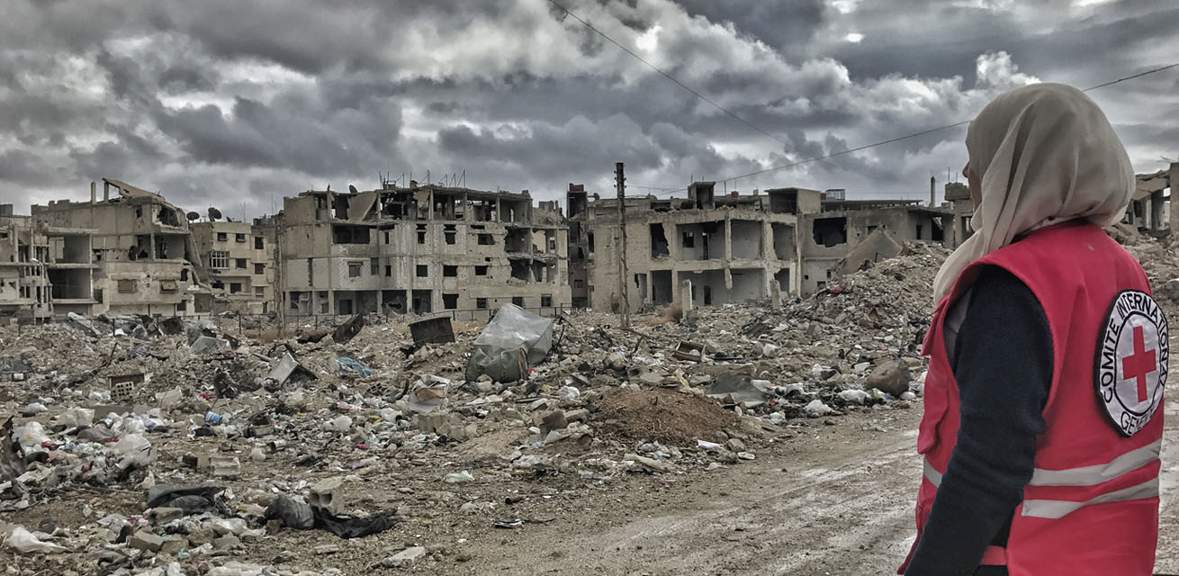 A town that has been destroyed by war, with a woman on the right in a Red Cross jacket looking at the rubble.
